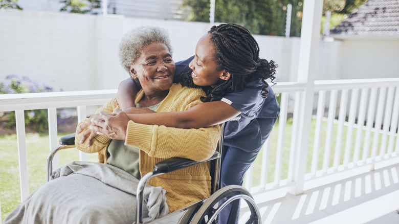 Older woman in a wheelchair being hugged from behind by woman wearing scrubs.