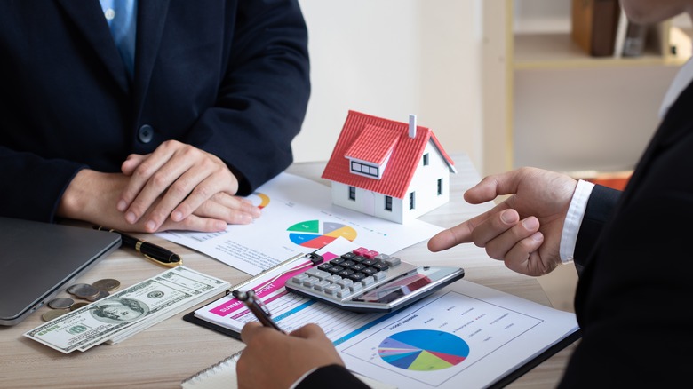 A table with paperwork of charts, a calculator, a model of a house and a pile of money with men's hands visible on either side  and one pointing to the calculator.