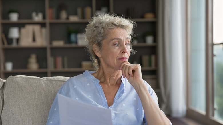 Pensive doubtful senior woman retiree sit on sofa hold business finance documents touch chin look away