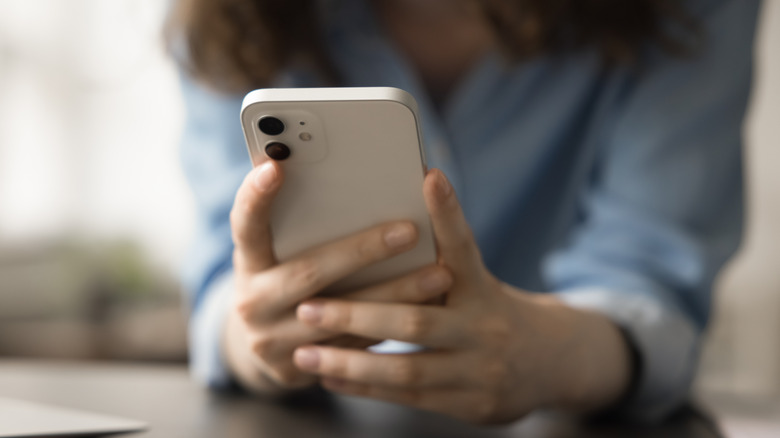 A woman holds a white smartphone, with phone in foreground, and woman blurred in background, in this shot that represents using a mobile app.